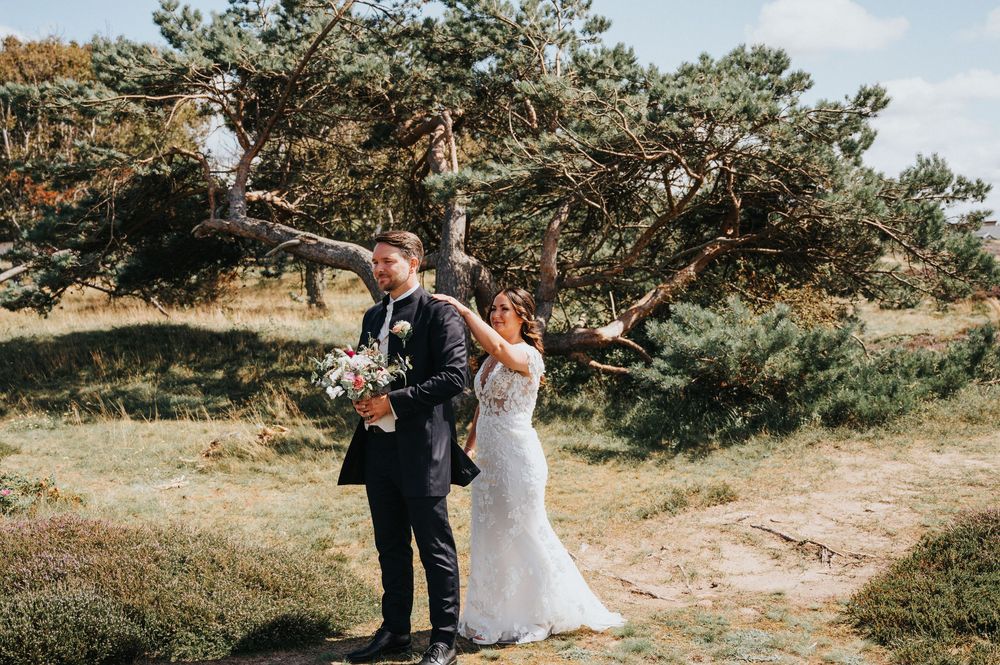 A couple in wedding attire stands together under a windswept tree on a scenic hillside path.