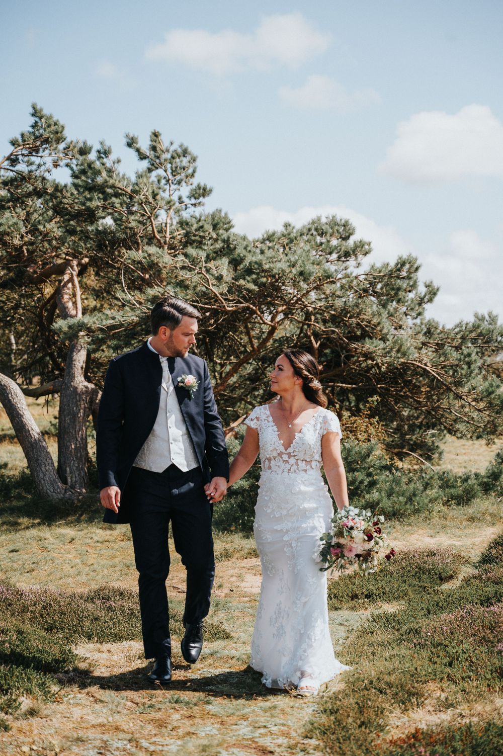 Newlywed couple walks together on a scenic hilltop path surrounded by pine trees on their wedding day.