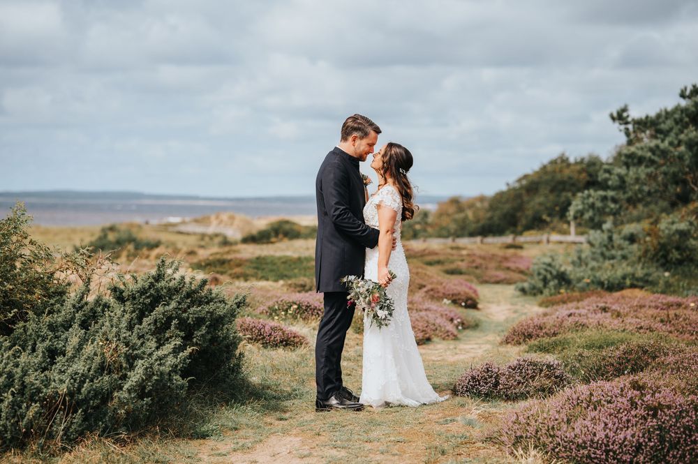 A couple shares a romantic moment on a heathland hilltop with purple heather and coastal views in the background.