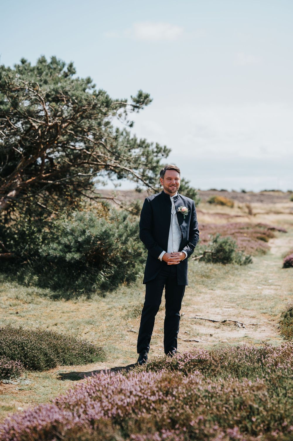 A person in a dark suit walks through a scenic heathland landscape with purple heather and pine trees.