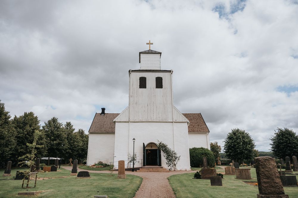 White church building with bell tower and cross stands on grassy grounds under cloudy sky with gravestones visible.