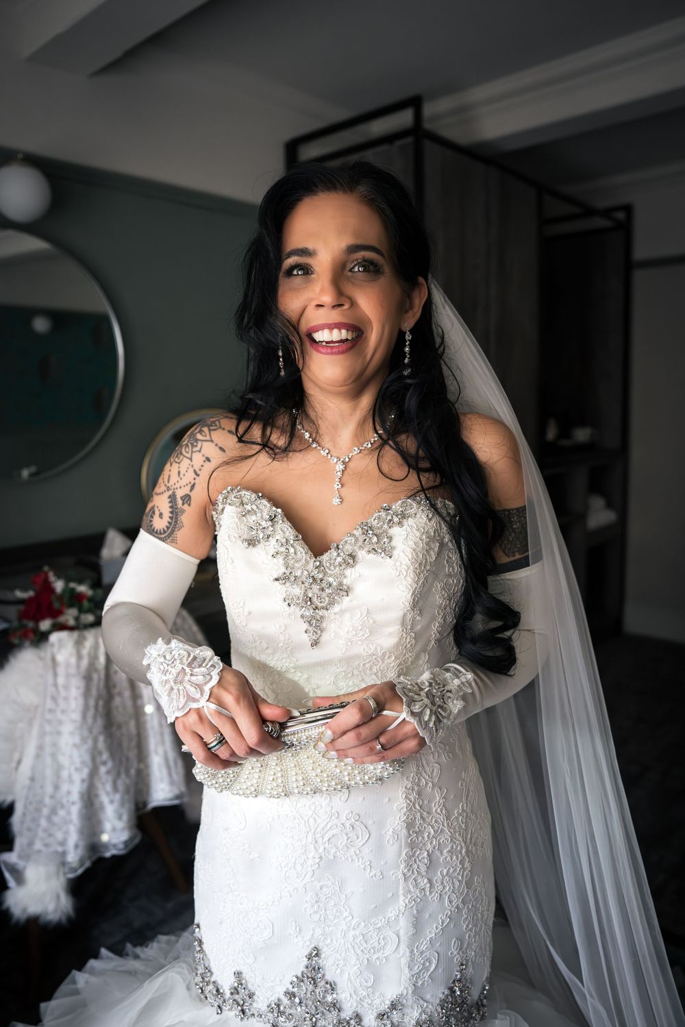 Smiling bride in strapless lace wedding gown with veil, silver jewelry, and tattoos on her arm.