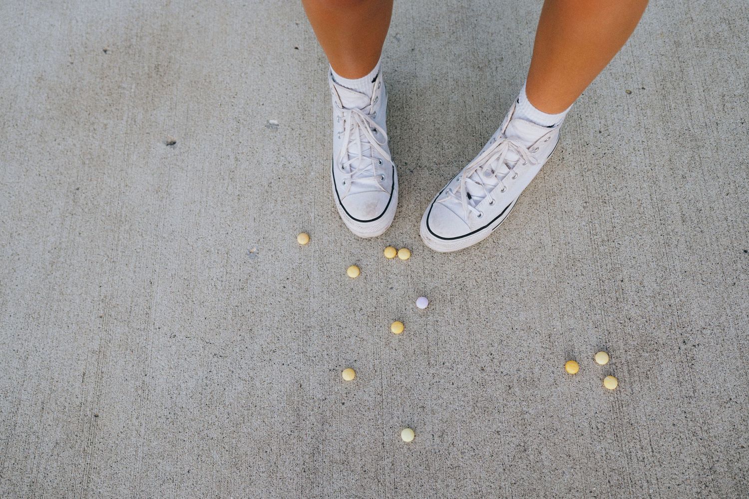 White sneakers and legs stand on gray concrete ground with scattered yellow and pink Mentos candy pieces.