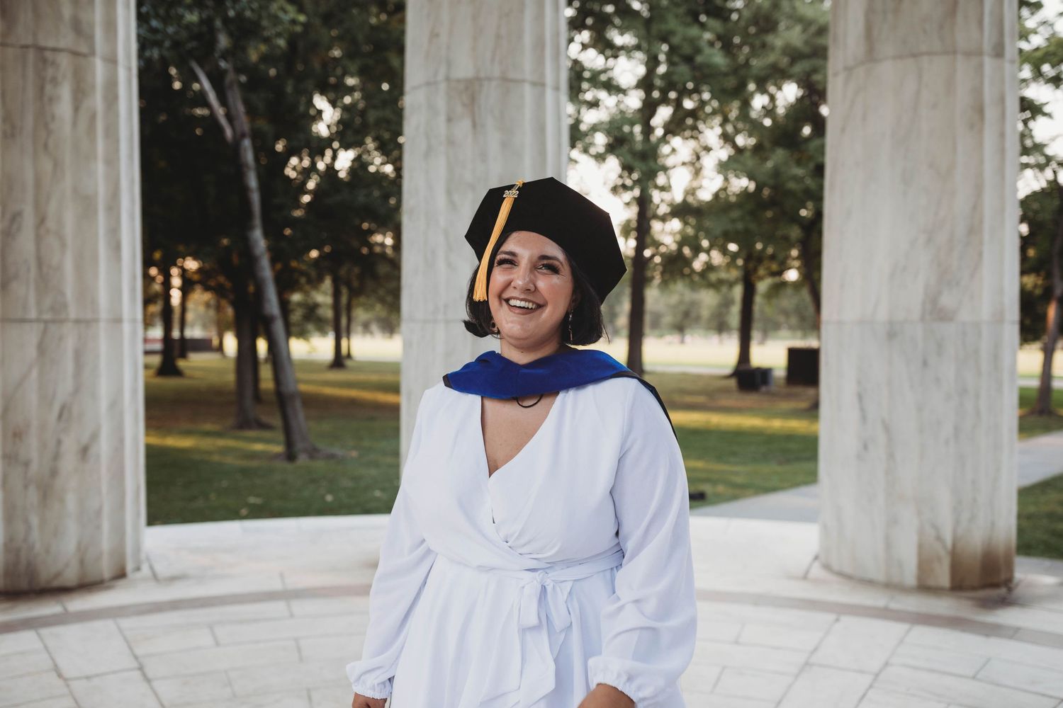 Proud graduate smiling in cap and gown standing between elegant white columns during outdoor graduation photo session.