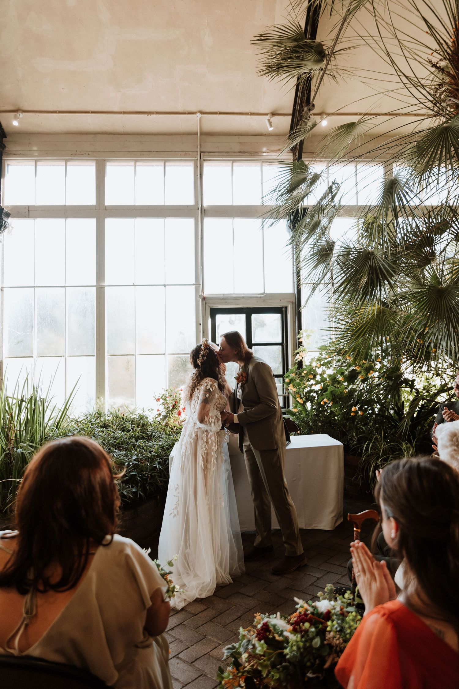 Wedding guests gather in a sunlit greenhouse venue decorated with plants and white dresses displayed along the windows.