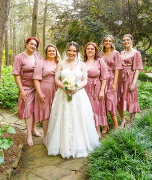 Wedding party wearing dusty rose pink dresses poses in a lush forest setting with white-gowned figure.