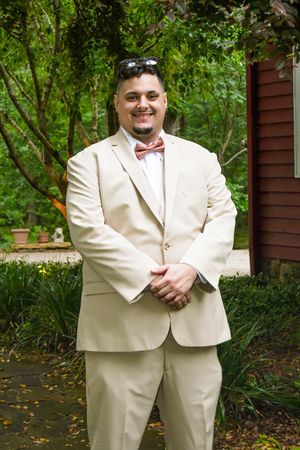 A person wearing a cream-colored suit and bowtie poses in a garden setting with lush greenery in the background.