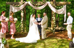 An outdoor wedding ceremony takes place under a wooden arch draped with white fabric in a lush wooded setting.