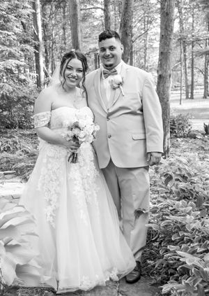 A bride and groom pose together in a wooded outdoor setting wearing white dress and light colored suit.
