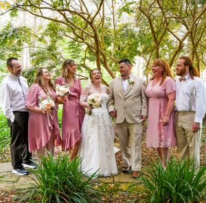 Wedding party stands in a garden setting with lush greenery and plants while wearing pink and neutral colored attire.