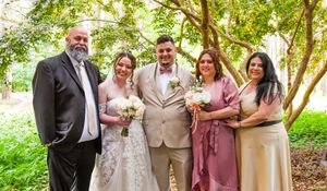 Wedding party in mauve and tan attire poses together under lush green trees during outdoor celebration.