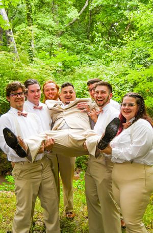 Groomsmen in khaki pants and white shirts pose playfully in a line against a lush green forest backdrop.