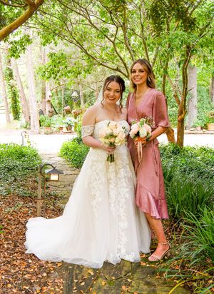 Two people in formal attire pose together outdoors, one wearing a white lace wedding gown and one in a dusty rose dress.