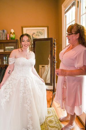 Wedding preparations showing a white lace dress and people in pink bridesmaid dresses in a sunlit room.