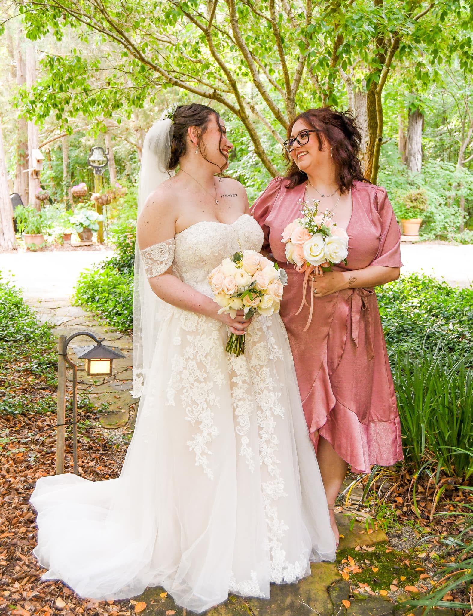 Bridesmaids in dusty rose dresses and bride in white lace gown pose together in a garden setting.