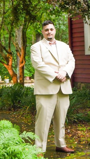 A person in a beige suit stands in a lush garden setting with green ferns and trees in the background.