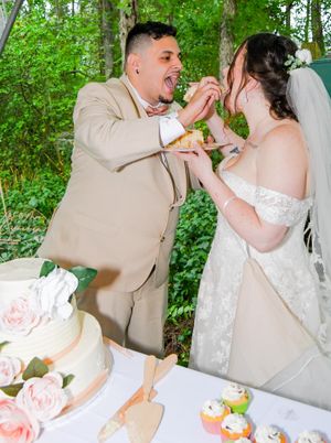 Wedding couple shares a playful cake-feeding moment in an outdoor garden setting with lush greenery.