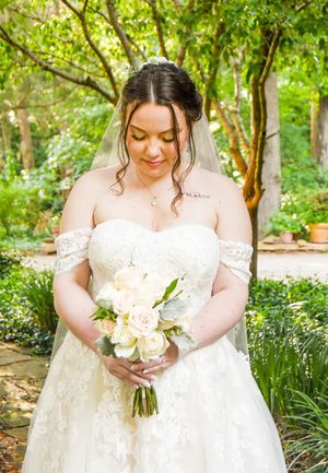 A bride in a white lace gown holds a delicate bouquet of blush roses in a lush garden setting.