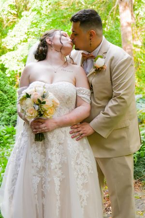 A romantic wedding couple shares tender kisses in a lush green garden setting while holding a delicate floral bouquet.