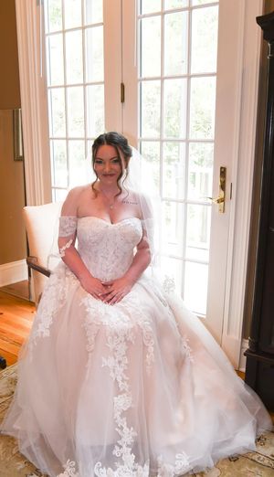 A bride in a stunning off-shoulder white lace ballgown poses by French doors in natural lighting.