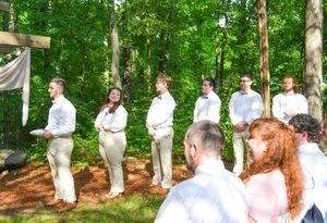 Wedding party members in white attire stand in line along a wooded path during an outdoor ceremony.
