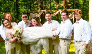 Wedding party wearing white poses playfully in a lush green forest setting during summer celebration.