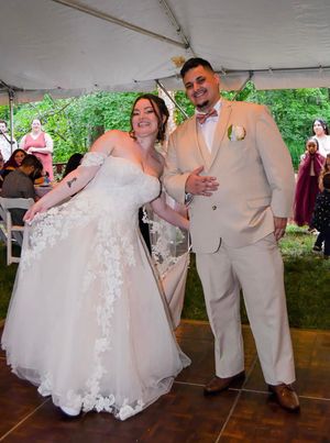 A couple in wedding attire dance together under a white tent, with the bride in a lace gown and groom in tan suit.