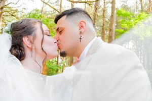 A couple in white wedding attire shares a tender moment in a sunlit forest setting.