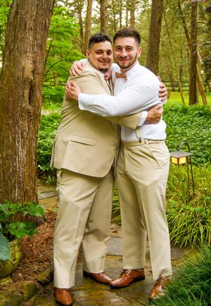 Wedding party members in beige suits share joyful embraces in a wooded outdoor setting.