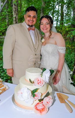 A couple cuts their white wedding cake decorated with pink roses during their outdoor forest reception.