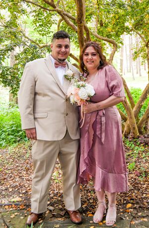 A couple poses outdoors in formal attire with the person in a beige suit and bow tie next to someone in a mauve dress.