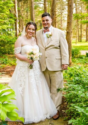 A wedding couple poses on a garden path surrounded by lush greenery wearing a white dress and tan suit.