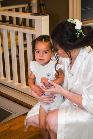 A tender moment between bride and child in white dresses sitting on wooden stairs during a wedding ceremony.