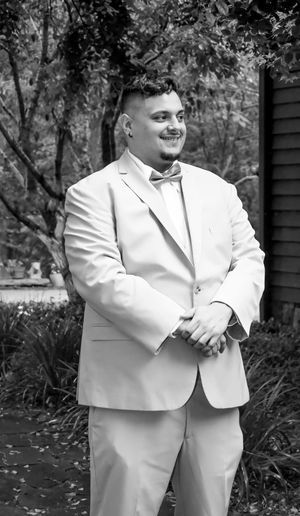 A person wearing a light colored suit and bow tie stands outdoors in a black and white photograph.