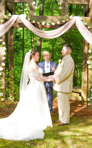 A romantic outdoor wedding ceremony takes place under a wooden pergola decorated with white fabric and string lights in a forest.