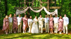 Wedding party members in coordinating pink and white attire pose together on grassy lawn for outdoor group photo.