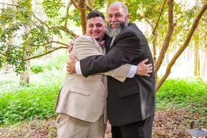 Two people embrace warmly in a lush green outdoor garden setting on a sunny day.