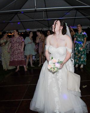 A bride in a white lace wedding dress stands holding a bouquet during a wedding reception.