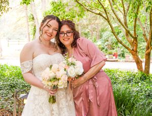 A group in formal attire poses together in a lush garden setting with pink and white floral accents.