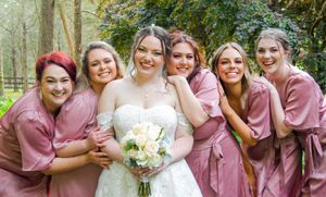 Bridesmaids wearing dusty rose dresses pose with bride holding white bouquet in outdoor wedding setting.