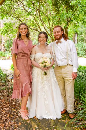 Wedding party poses outdoors in mauve bridesmaid dresses and white gown against lush green garden backdrop.