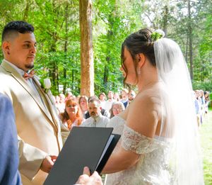 An outdoor wedding ceremony takes place in a wooded setting with guests gathered around.