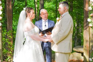 An intimate wedding ceremony unfolds beneath a rustic wooden arbor adorned with white draping in a lush green forest.
