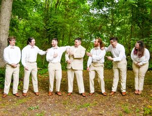 A wedding party wearing all white outfits stands in a line against a lush green forest backdrop.