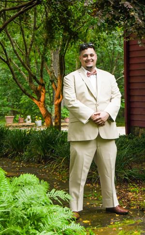 Person in a cream-colored suit poses in a garden setting with lush green ferns and trees in the background.