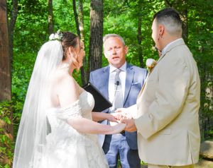 A wedding ceremony takes place in a lush green forest setting with sunlight filtering through the trees.