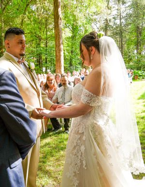An intimate outdoor wedding ceremony in a wooded setting with guests watching the couple exchange rings.