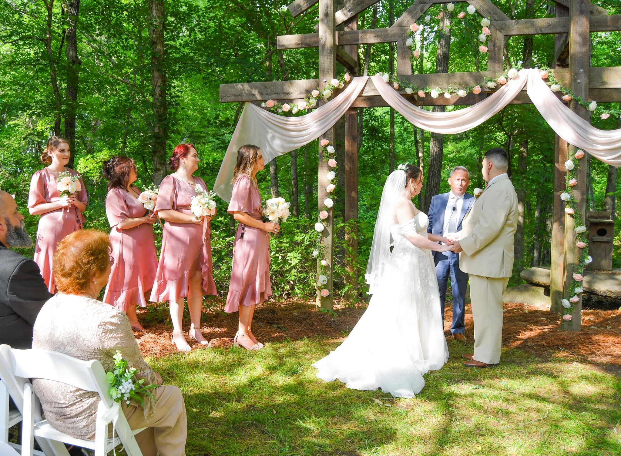 A panoramic view of an outdoor wedding ceremony setup with pink decor and white chairs in a forest setting.