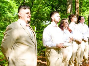 Wedding party members in tan and white attire stand together in a lush green forest setting.