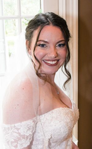 A white lace off-shoulder wedding dress is showcased near a bright window with natural lighting.